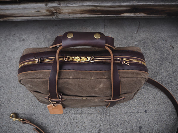 Top of light brown canvas briefcase with dark brown leather handles.  Antique brass zipper and rivets.