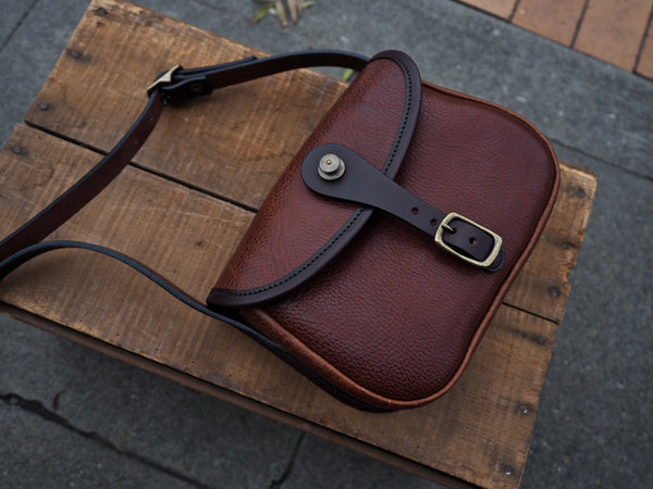 Brown leather purse with flap and dark brown leather binding. Solid brass quick snap and rounded buckle.  Dark brown leather shoulder strap.