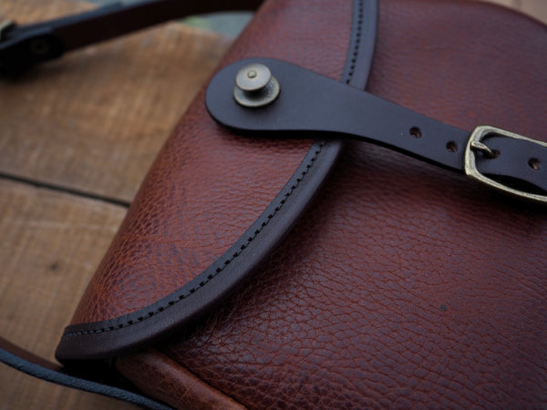 Brown leather purse with flap and dark brown leather binding. Solid brass quick snap and rounded buckle.  Dark brown leather shoulder strap.