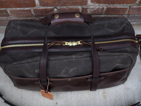 Top of dark green canvas bag has brass YKK zipper.  Dark brown handle wrap holds together dark brown handles.