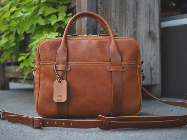 Light brown leather briefcase measuring 15.5 inches long, 11.75 inches tall and 4.5 inches wide. Two light brown leather straps riveted to front leather pocket that turn into rolled handles. Light brown leather shoulder strap with antique brass roller buckles attached to briefcase.