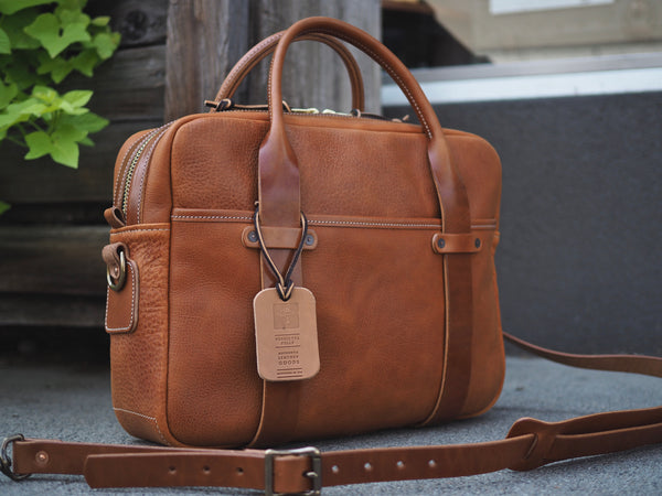 Light brown leather briefcase measuring 15.5 inches long, 11.75 inches tall and 4.5 inches wide. Two light brown leather straps riveted to front leather pocket that turn into rolled handles. Light brown leather shoulder strap with antique brass roller buckles attached to briefcase.